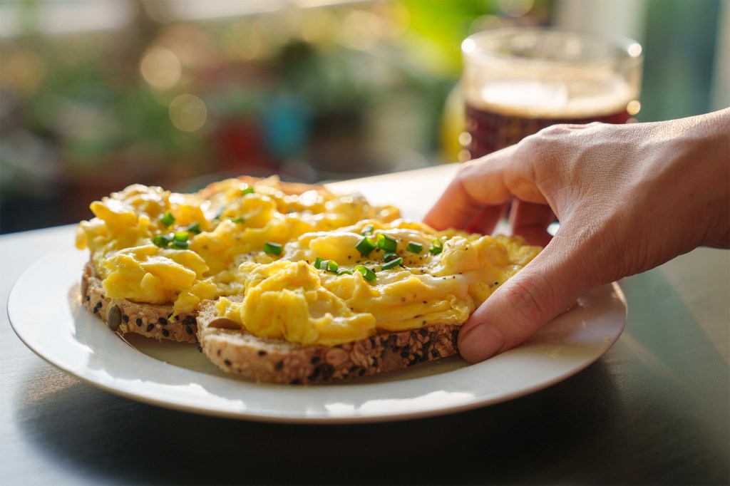 A hand holding a plate of soft scrambled eggs on toast